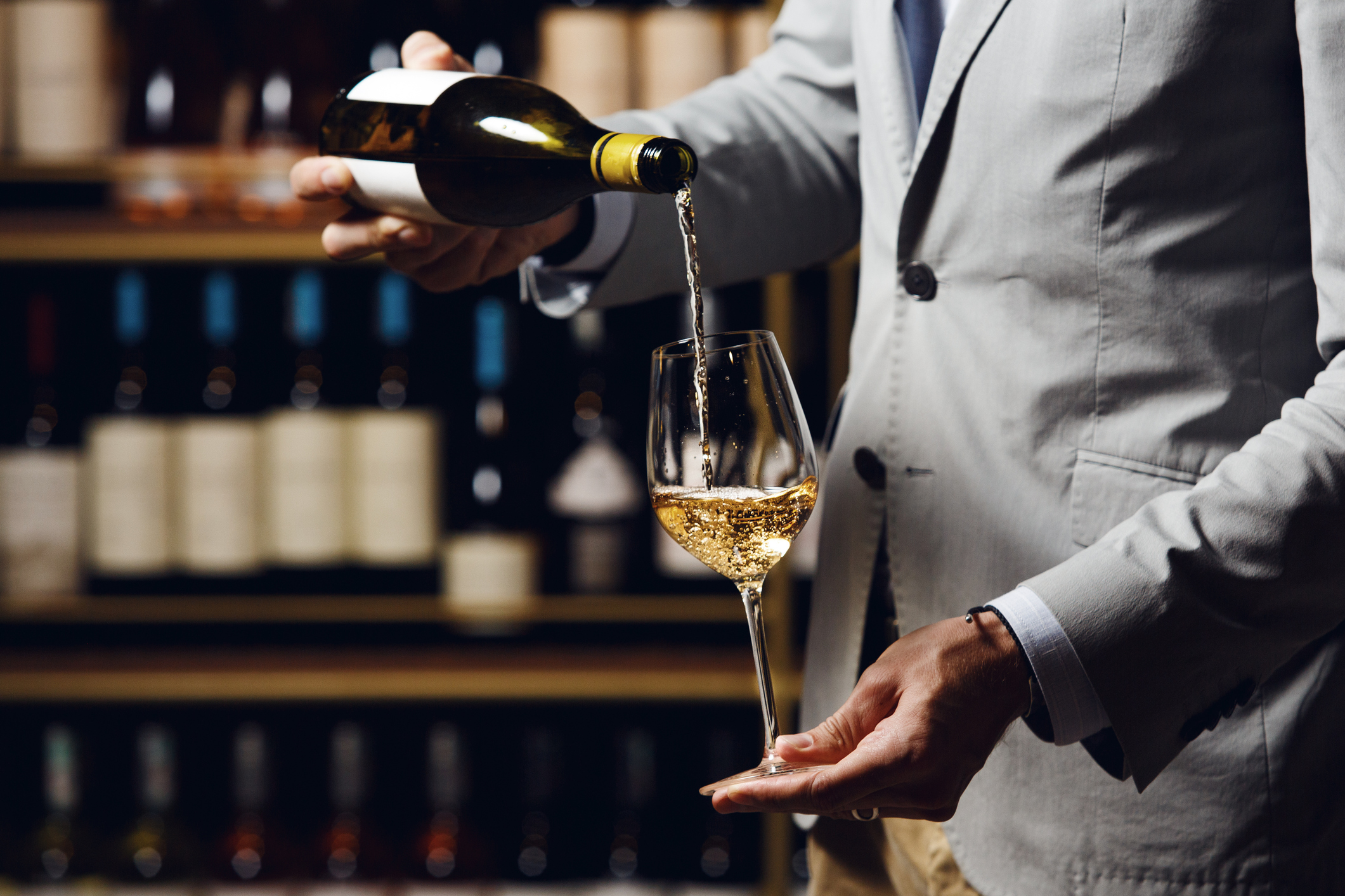 Close up indoor portrait of male sommelier pouring white wine into long-stemmed wineglass. Waiter with bottle of alcohol beverage iwth a free copy space. Bartender in cellar pour elite drink standing against wine shelf