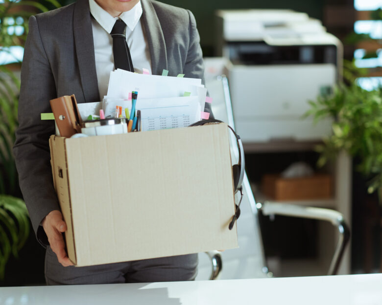 New job. Closeup on modern female employee in modern green office in grey business suit with personal belongings in cardboard box.