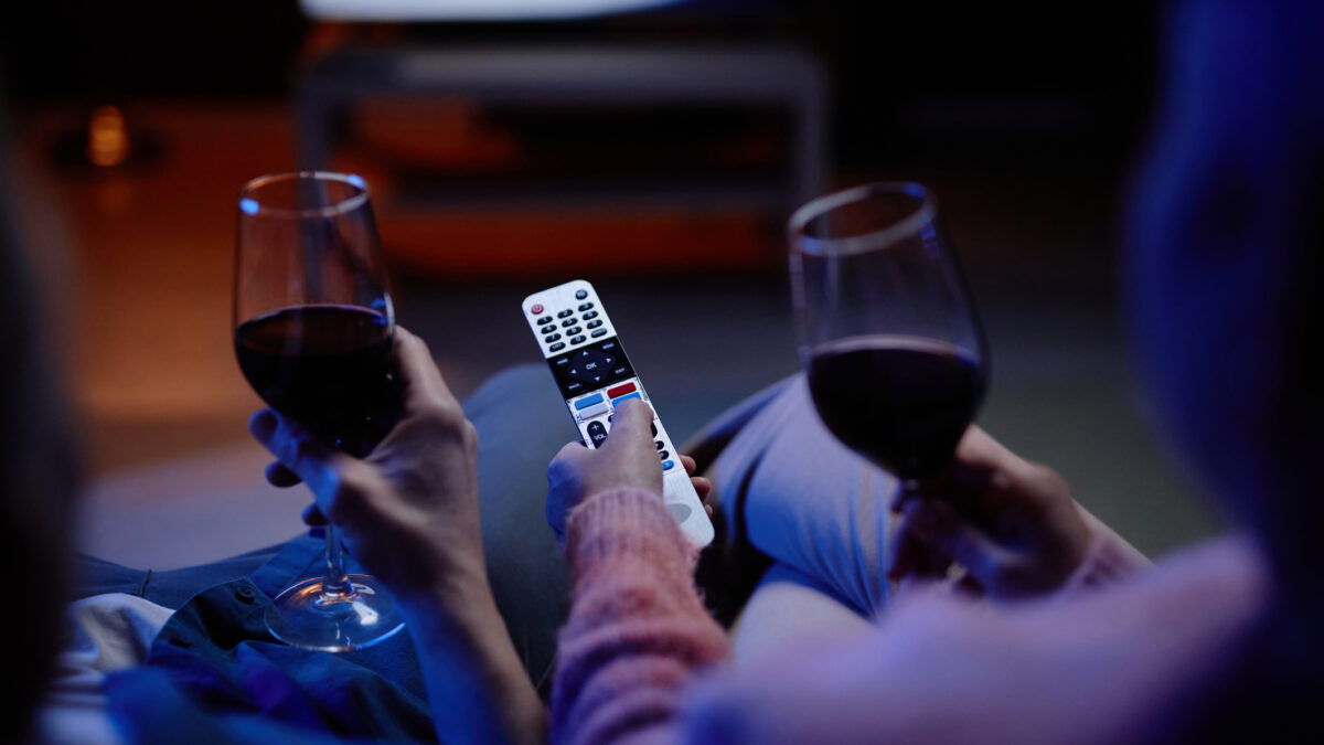 Top view closeup of unrecognizable senior couple watching TV together with hands holding remote control and wine glasses
