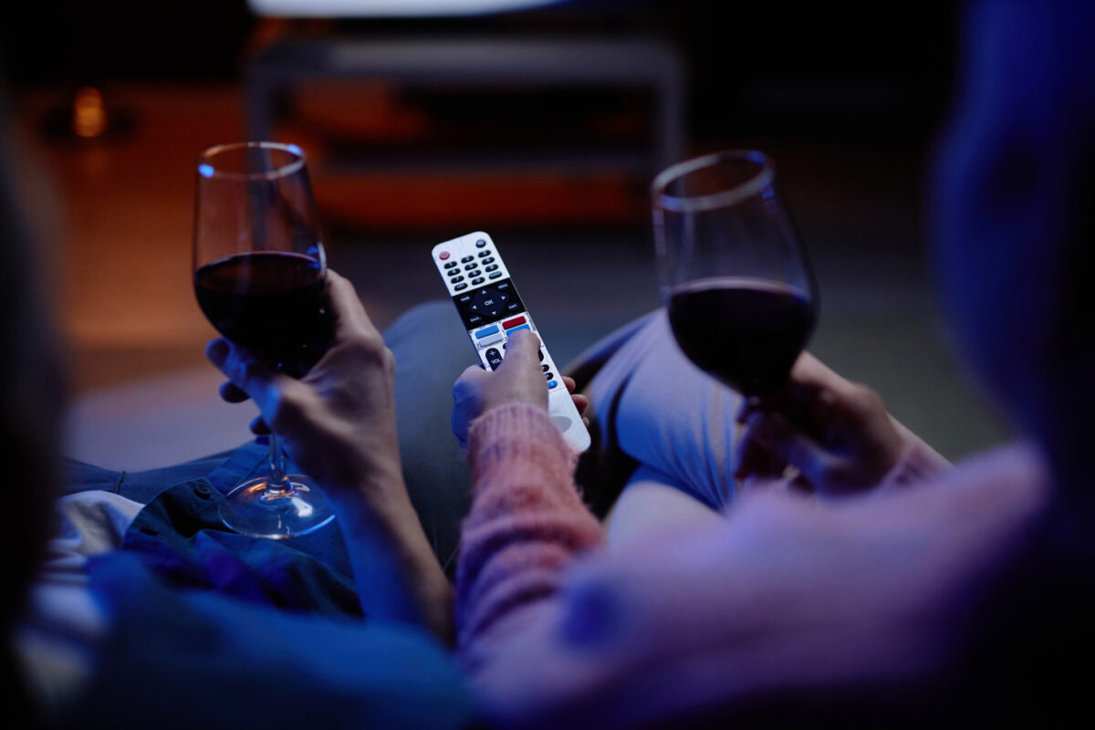 Top view closeup of unrecognizable senior couple watching TV together with hands holding remote control and wine glasses