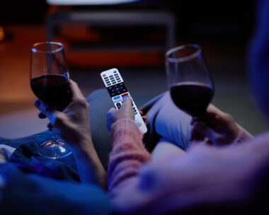 Top view closeup of unrecognizable senior couple watching TV together with hands holding remote control and wine glasses
