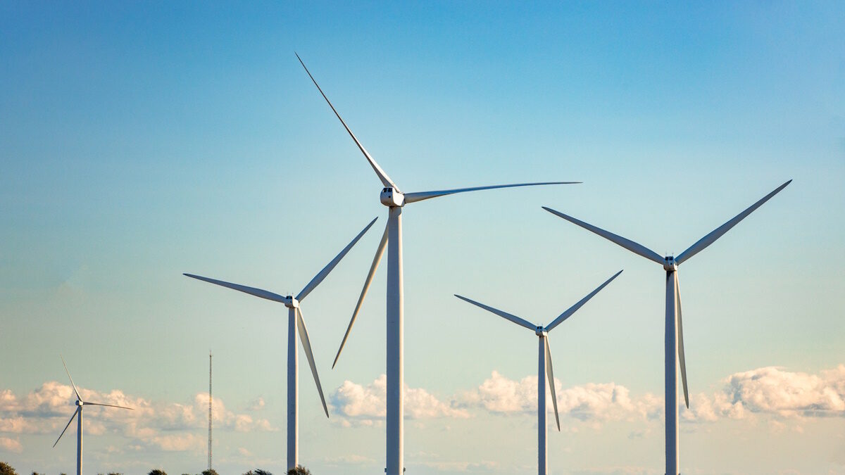 Color image depicting many wind turbines in a row on a wind farm nestled within an agricultural landscape.