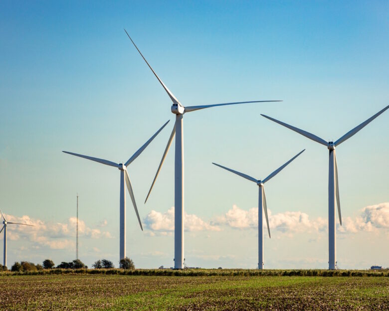Color image depicting many wind turbines in a row on a wind farm nestled within an agricultural landscape.