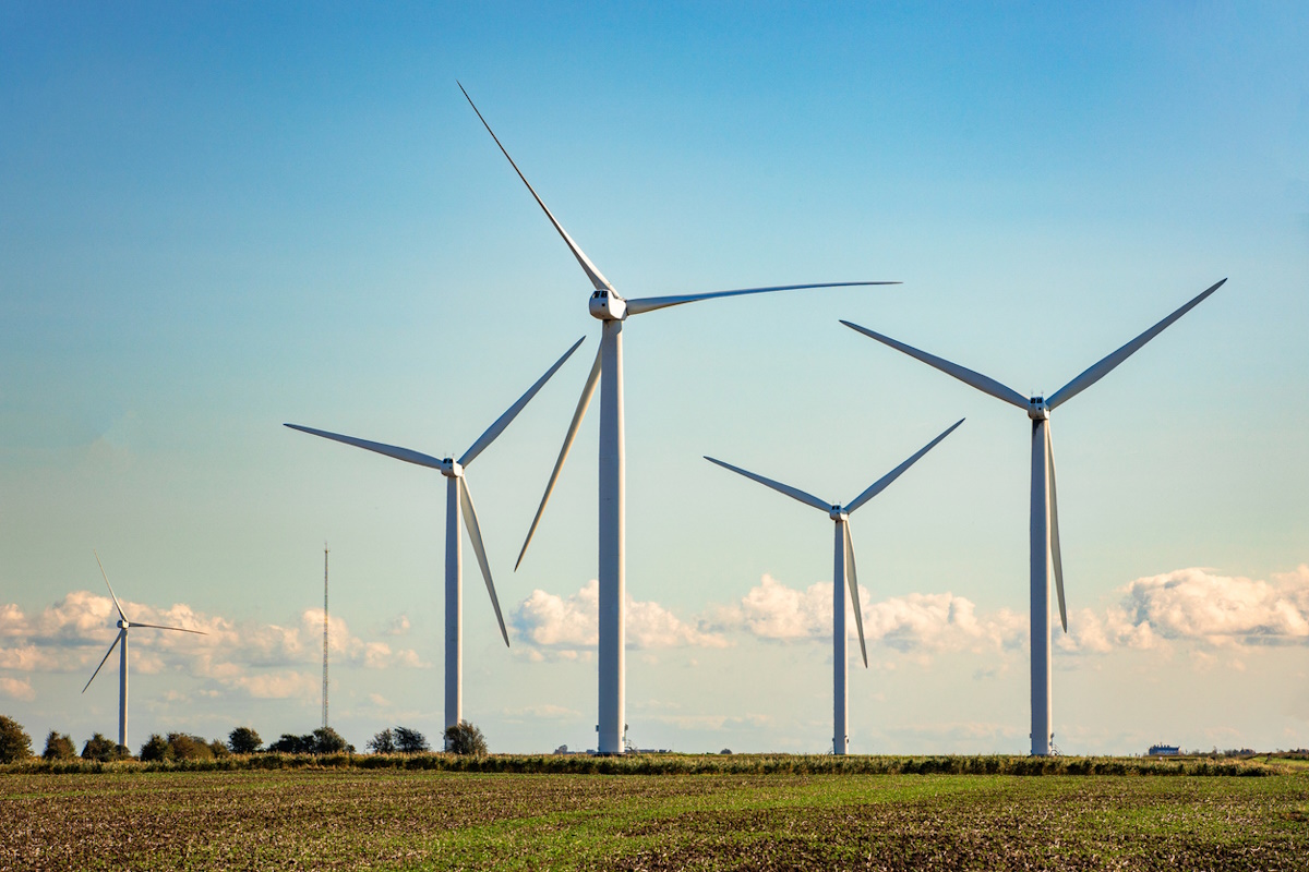 Color image depicting many wind turbines in a row on a wind farm nestled within an agricultural landscape.