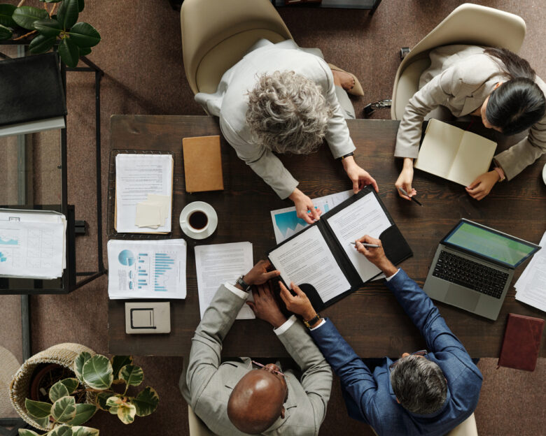Group of middle aged multiethnic business professionals collaborating around table, reviewing documents and using laptop, top view showing teamwork and corporate meeting environment