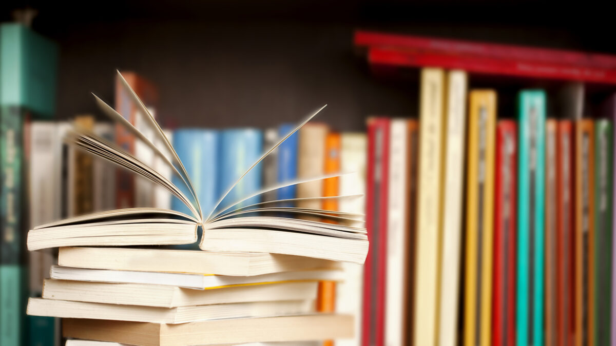 Stack of books on a wooden library shelf, one of them open on top, multicolored book spines background.