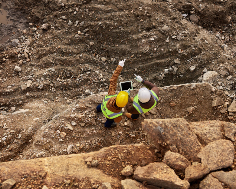 Top view shot of two industrial workers wearing reflective jackets standing on mining worksite outdoors using digital tablet, copy space
