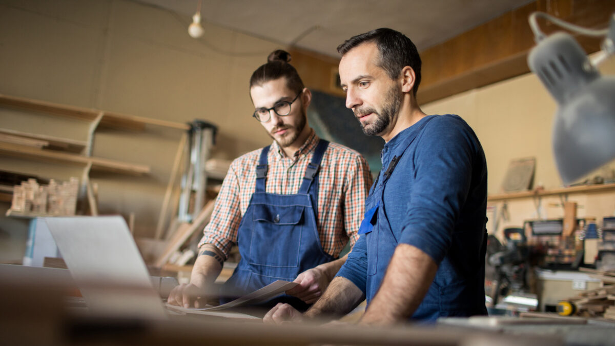 Low angle portrait of two modern artisans using laptop in workshop while working with wood together, copy space