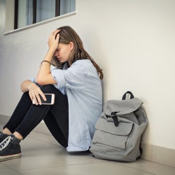 Upset and depressed girl holding smartphone sitting on college campus floor holding head. University sad student suffering from depression sitting on floor at high school. Lonely bullied teen in difficulty with copy space.