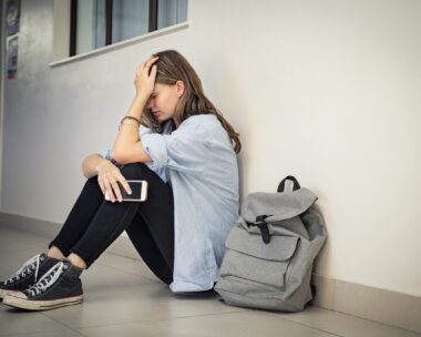 Upset and depressed girl holding smartphone sitting on college campus floor holding head. University sad student suffering from depression sitting on floor at high school. Lonely bullied teen in difficulty with copy space.