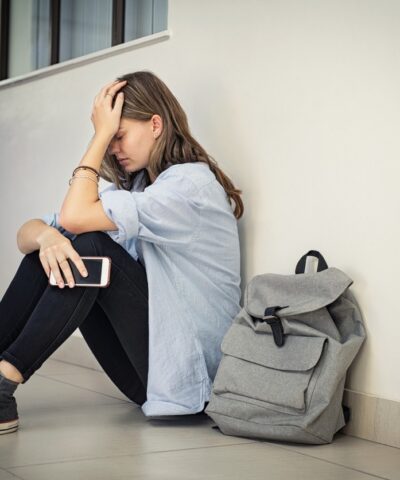Upset and depressed girl holding smartphone sitting on college campus floor holding head. University sad student suffering from depression sitting on floor at high school. Lonely bullied teen in difficulty with copy space.
