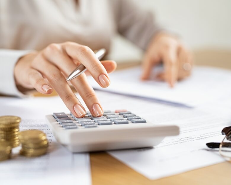 Woman using a calculator doing a balance of monthly expenses. Close up of businesswoman working with financial data and using calculator with coins on desk at office. Deatil of hands calculating home finances and annual taxes.