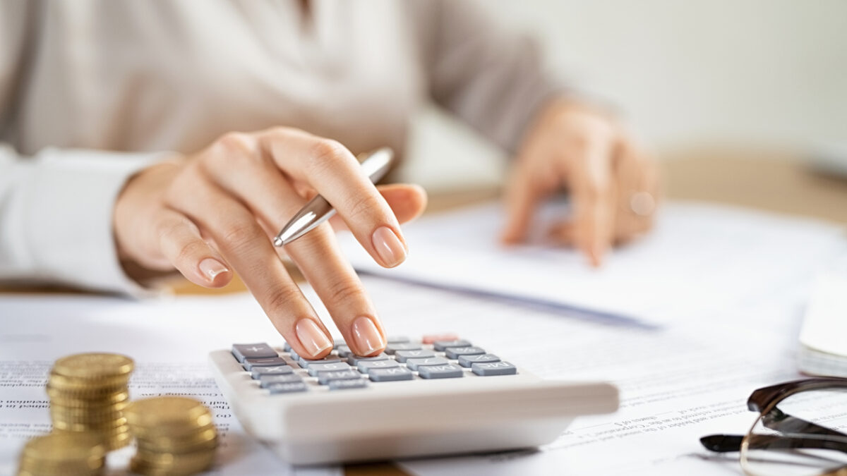 Woman using a calculator doing a balance of monthly expenses. Close up of businesswoman working with financial data and using calculator with coins on desk at office. Deatil of hands calculating home finances and annual taxes.