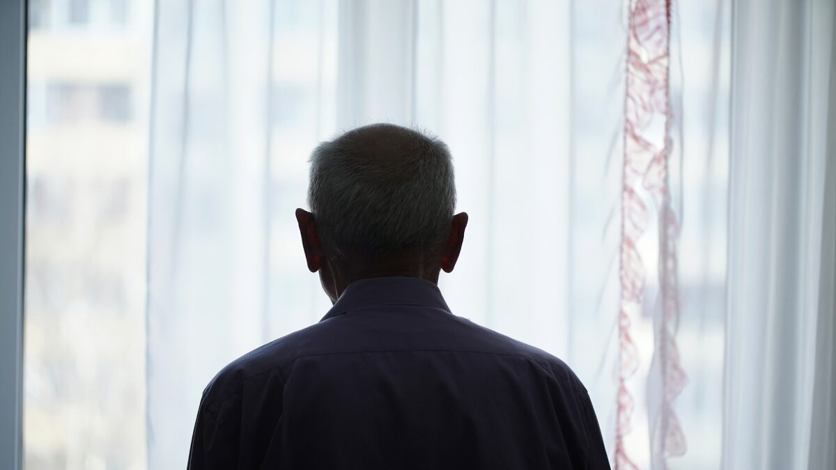 Silhouette of retired man looking through window with transparent curtain standing at home rear view. Loneliness and old human care concept