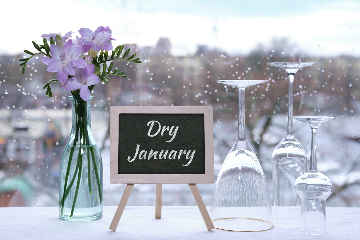 Dry January, month without alcohol. Text Dry January on blackboard, chalk board. Empty vine and beer drinking glasses, freesia flowers. Window with raindrops, grey winter city skyline on background.