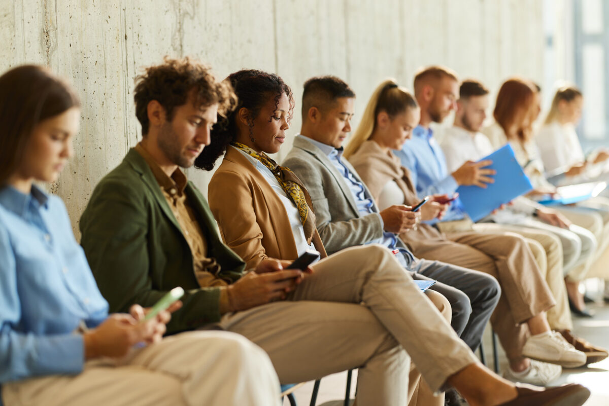 Large group of business people waiting for a job interview in a hallway. Focus is on black woman using mobile phone.
