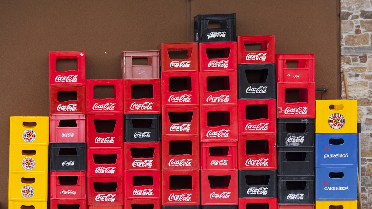 Group of stacks of multi colored plastic crates for beverage bottles, seen on a town street sidewalk outside a bar. Several beer, soda and soft drink brands on them. Concrete wall in the background.