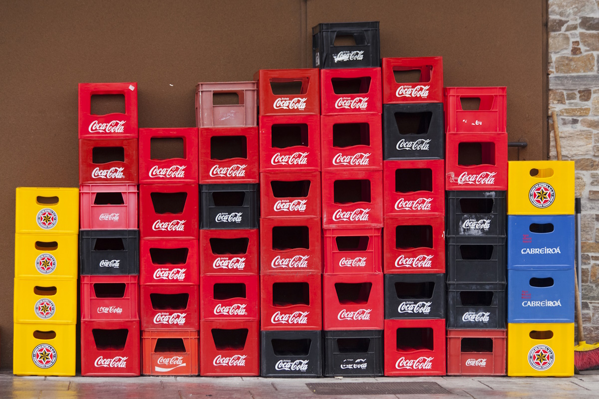Group of stacks of multi colored plastic crates for beverage bottles, seen on a town street sidewalk outside a bar. Several beer, soda and soft drink brands on them. Concrete wall in the background.