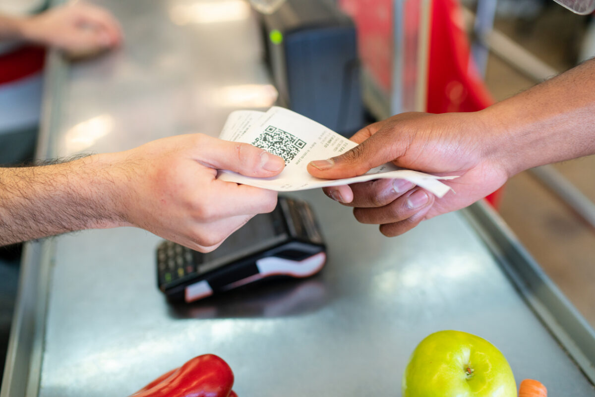 Hands exchange a receipt at a supermarket checkout counter after a grocery purchase. Two people finalize a transaction exchanging a paper slip containing a QR code at grocery store checkout counter