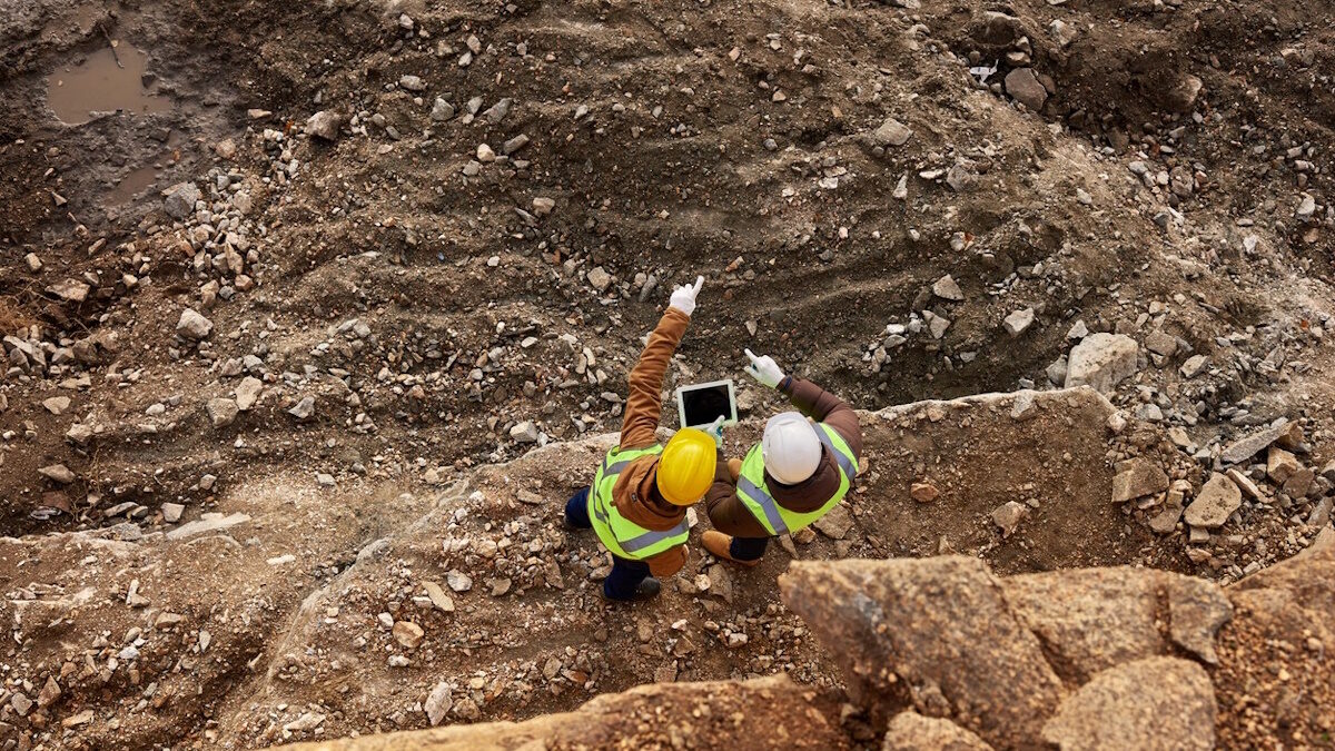 Top view shot of two industrial workers wearing reflective jackets standing on mining worksite outdoors using digital tablet, copy space