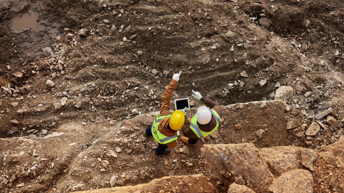 Top view shot of two industrial workers wearing reflective jackets standing on mining worksite outdoors using digital tablet, copy space