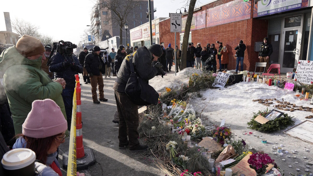 Στέλεχος του Γραφείου Ποινικής Σύλληψης (BCA) επιμελείται το σημείο όπου ο Alex Pretti, σκοτώθηκε από πυροβολισμούς ομοσπονδιακών υπαλλήλων της υπηρεσίας μετανάστευσης που προσπάθησαν να τον συλλάβουν, ενώ άνθρωποι συγκεντρώνονται σε ένα αυτοσχέδιο μνημείο, στο Μινεάπολις