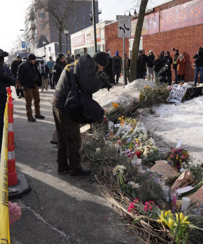 Στέλεχος του Γραφείου Ποινικής Σύλληψης (BCA) επιμελείται το σημείο όπου ο Alex Pretti, σκοτώθηκε από πυροβολισμούς ομοσπονδιακών υπαλλήλων της υπηρεσίας μετανάστευσης που προσπάθησαν να τον συλλάβουν, ενώ άνθρωποι συγκεντρώνονται σε ένα αυτοσχέδιο μνημείο, στο Μινεάπολις