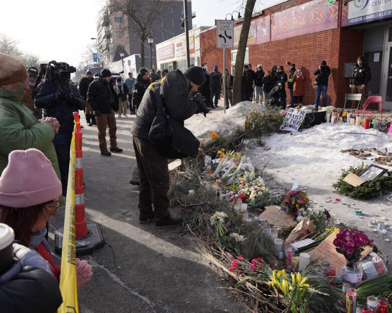 Στέλεχος του Γραφείου Ποινικής Σύλληψης (BCA) επιμελείται το σημείο όπου ο Alex Pretti, σκοτώθηκε από πυροβολισμούς ομοσπονδιακών υπαλλήλων της υπηρεσίας μετανάστευσης που προσπάθησαν να τον συλλάβουν, ενώ άνθρωποι συγκεντρώνονται σε ένα αυτοσχέδιο μνημείο, στο Μινεάπολις