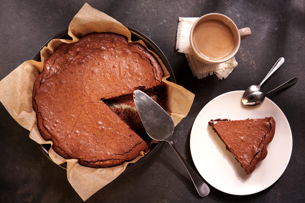 Homemade dark chocolate cake brownie in baking pan, mug with tea or coffee, chocolate on dark table with window light. Delicious bitter sweet and fudge. Flat lay, top view, vintage style.