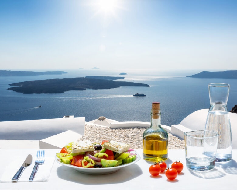 A Greek salad with olive oil and tomatoes in front of the a breathtaking view to the blue, Aegean sea as a concept of Greek food in summer time