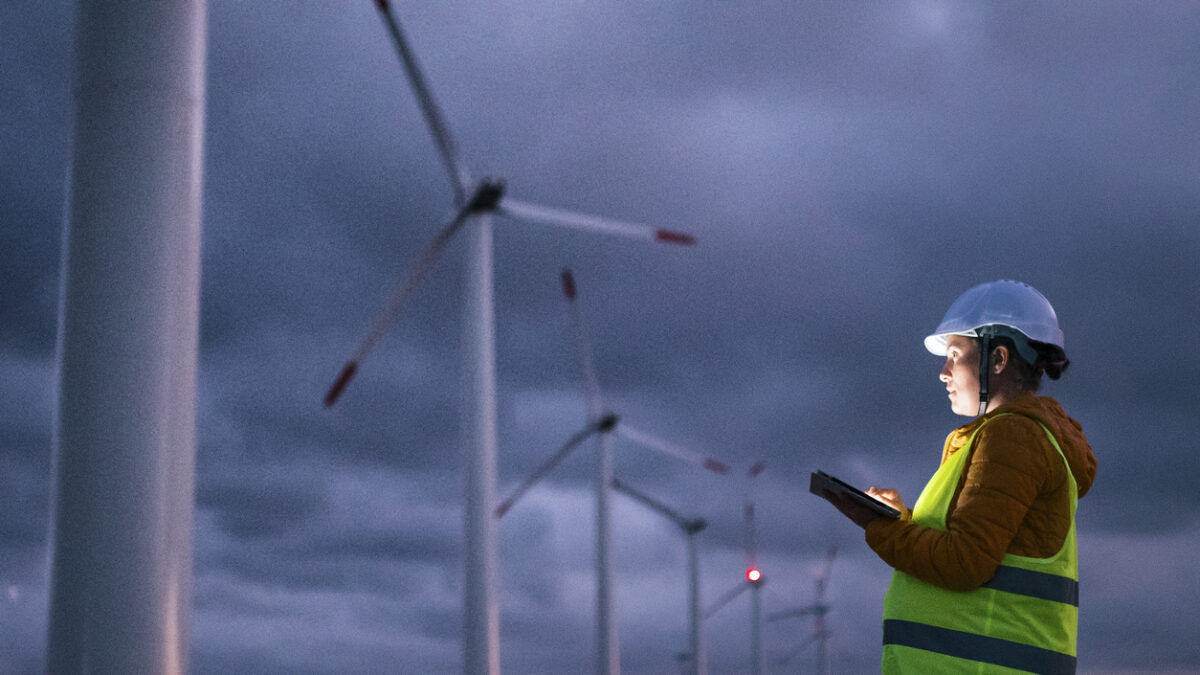 Electrical engineer working for the energy industry, supervising the condition of the Electrical Power Equipment in a wind turbines farm power station at night. Checking the data and the results of measurements with digital tablet. Pregnant woman engineer working on the field. Technology and Global Business.