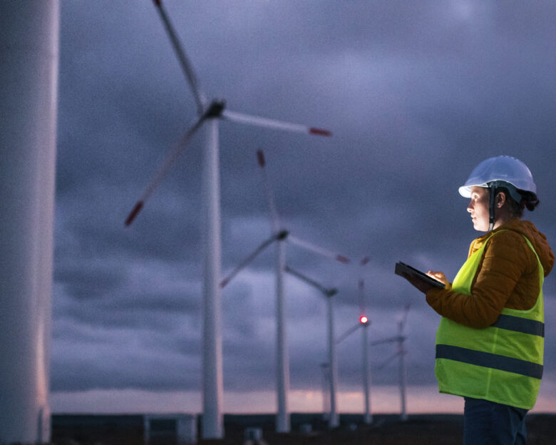 Electrical engineer working for the energy industry, supervising the condition of the Electrical Power Equipment in a wind turbines farm power station at night. Checking the data and the results of measurements with digital tablet. Pregnant woman engineer working on the field. Technology and Global Business.