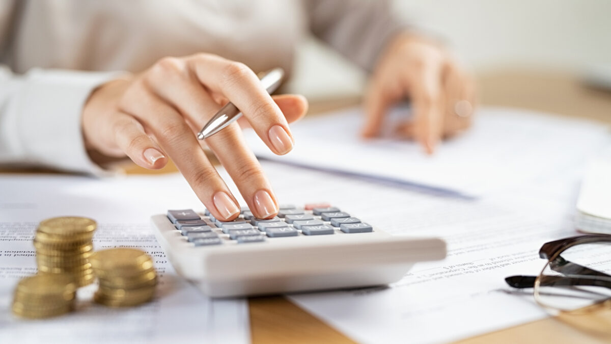 Woman using a calculator doing a balance of monthly expenses. Close up of businesswoman working with financial data and using calculator with coins on desk at office. Deatil of hands calculating home finances and annual taxes.