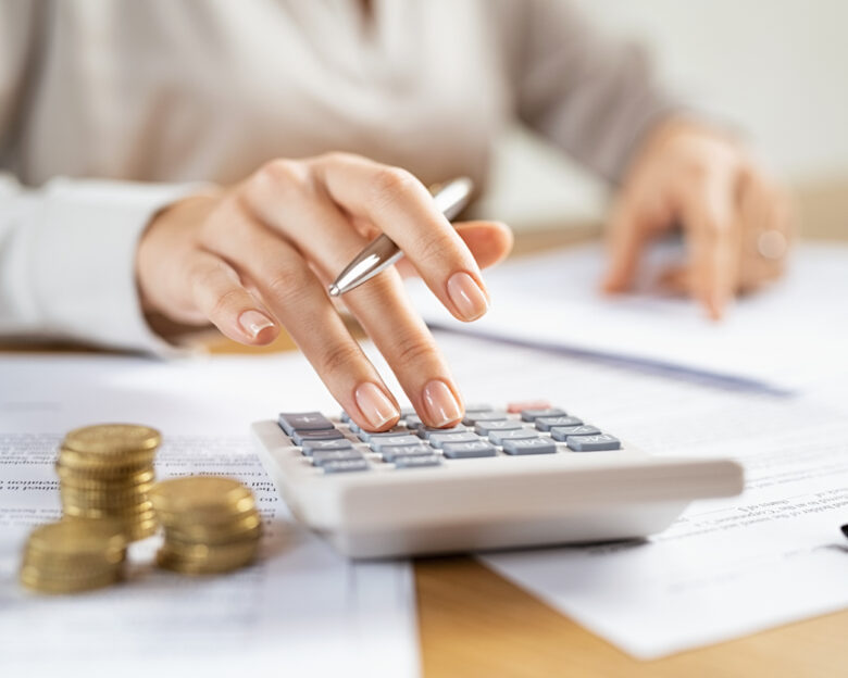 Woman using a calculator doing a balance of monthly expenses. Close up of businesswoman working with financial data and using calculator with coins on desk at office. Deatil of hands calculating home finances and annual taxes.