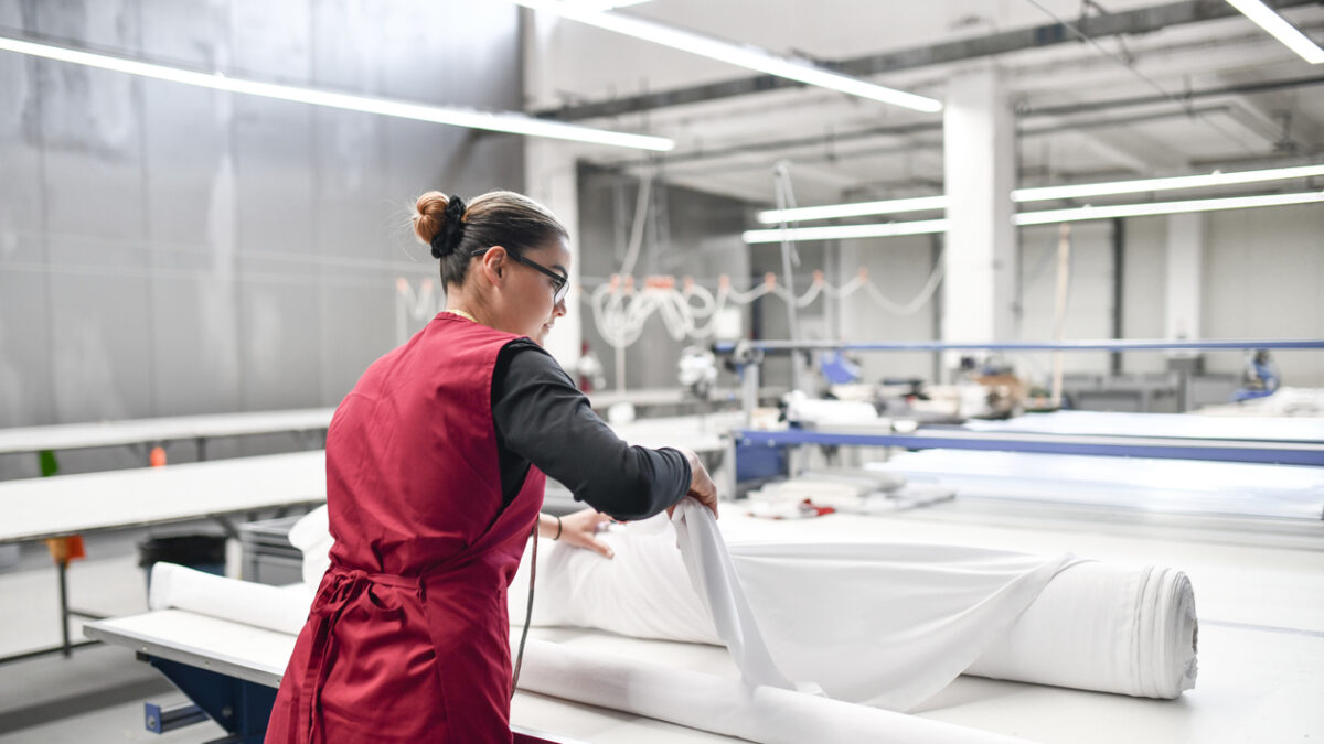 Rear View Of Expert Female Textile Worker Unrolling Material Rolls For Cutting