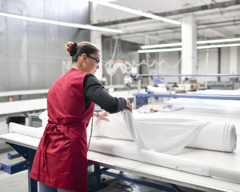 Rear View Of Expert Female Textile Worker Unrolling Material Rolls For Cutting