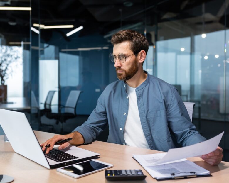 A serious young man accountant, financier, analyst, auditor sits in the office at the table. He holds documents and a pen in his hands, checks accounts, finances, types on a laptop.