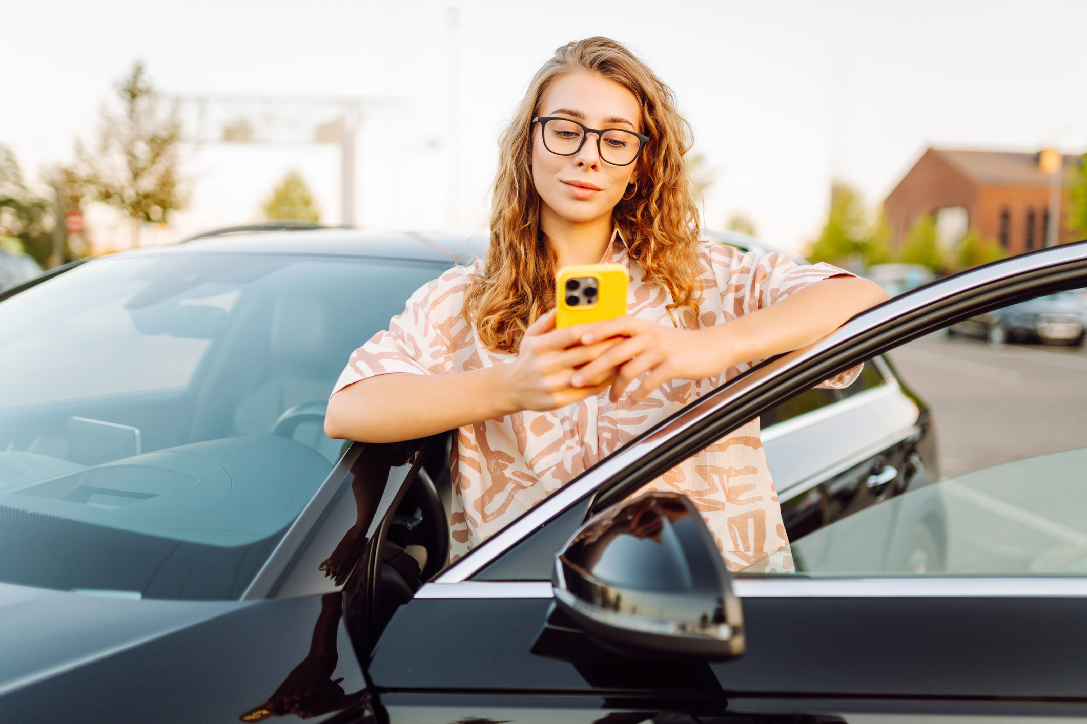 Young woman with glasses and a phone near her car in a parking lot. Beautiful woman with a phone, blogging at sunset outdoors. Concept of rest, relaxation.