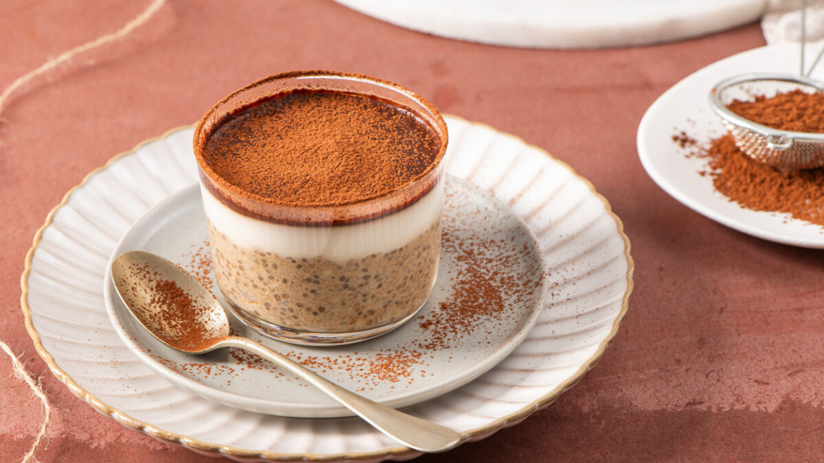 Tiramisu chia pudding in a glass cup on a white plate with a spoon, garnished with cocoa powder, set on a brown surface with a sieve and cocoa powder in the background