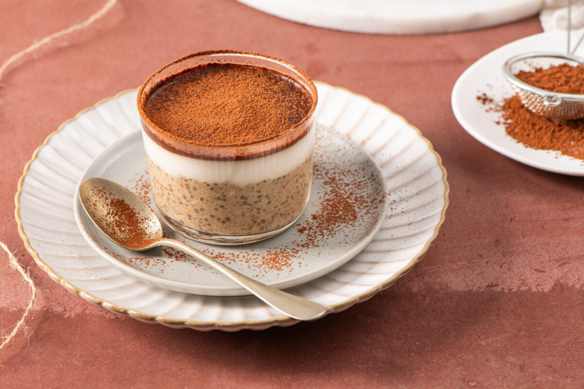 Tiramisu chia pudding in a glass cup on a white plate with a spoon, garnished with cocoa powder, set on a brown surface with a sieve and cocoa powder in the background