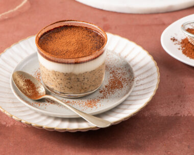 Tiramisu chia pudding in a glass cup on a white plate with a spoon, garnished with cocoa powder, set on a brown surface with a sieve and cocoa powder in the background