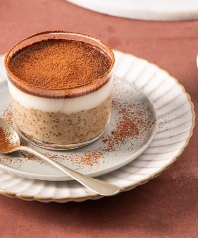 Tiramisu chia pudding in a glass cup on a white plate with a spoon, garnished with cocoa powder, set on a brown surface with a sieve and cocoa powder in the background