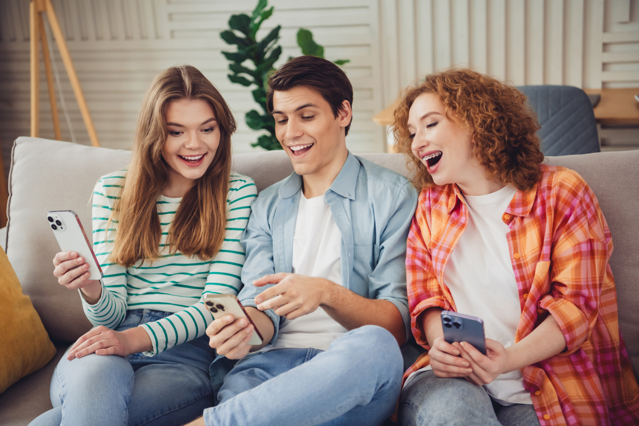Three cheerful friends relax on a cozy sofa, enjoying their time together, interacting with their smartphones and creating joyful moments in an inviting and comfortable living space.