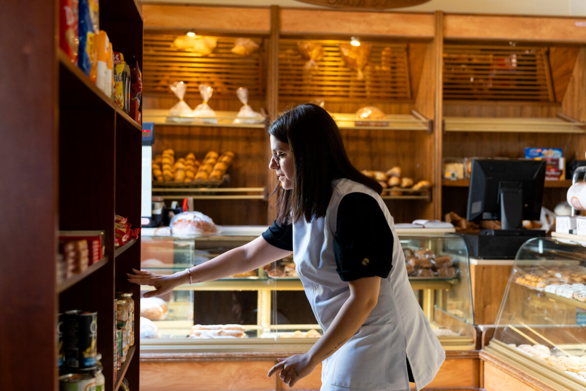 Woman in apron stocking shelves with bread, pastries and groceries in a small, family-run bakery shop, mid-adult owner at work