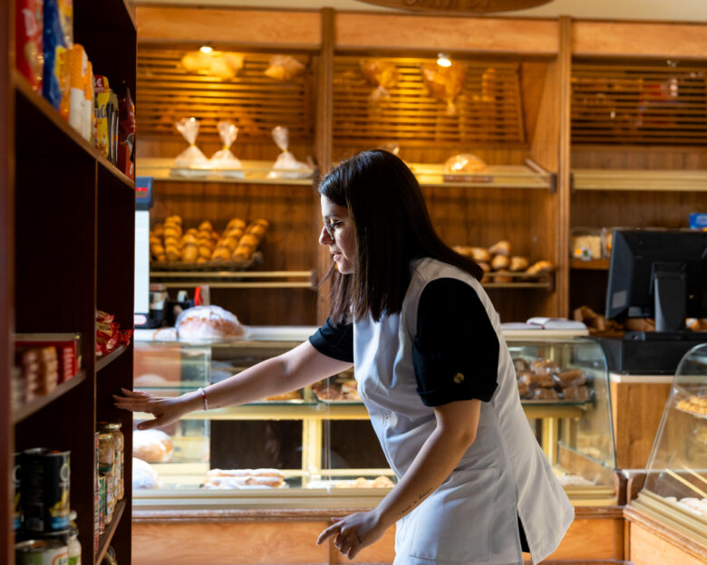 Woman in apron stocking shelves with bread, pastries and groceries in a small, family-run bakery shop, mid-adult owner at work