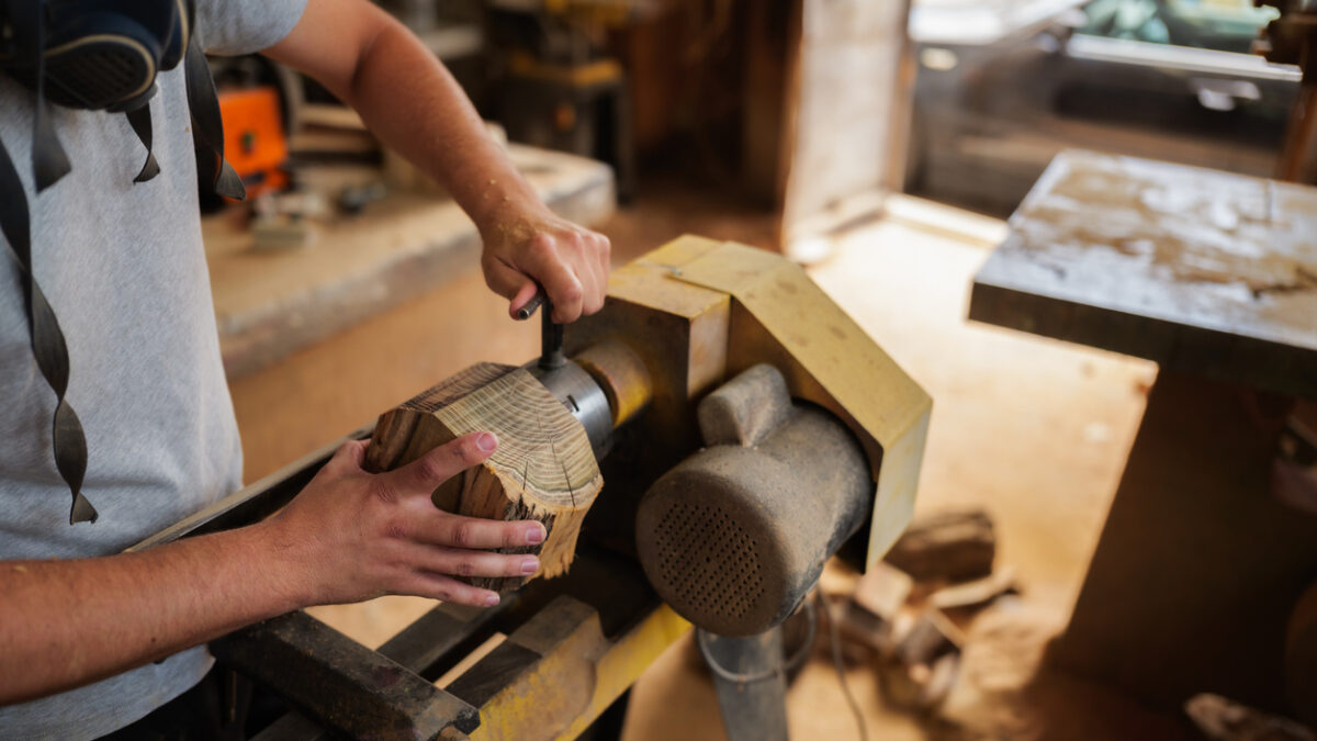 Carpenter wearing a protective mask, skillfully working wood on a lathe in an old, dusty workshop, meticulously crafting a unique handmade piece of furniture with precision and care