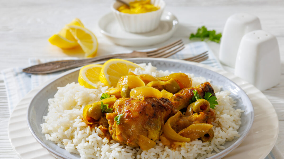 close-up of Chicken Yassa stewed in mustard, served with caramelized onions, olives, and basmati rice on a white plate on white wood table,close-up, not AI generated