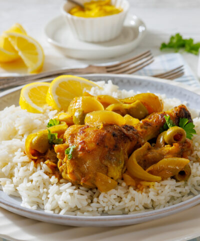 close-up of Chicken Yassa stewed in mustard, served with caramelized onions, olives, and basmati rice on a white plate on white wood table,close-up, not AI generated