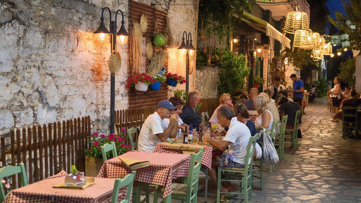 Pythagoreio is a small town and former municipality on the island of Samos, North Aegean, Greece. Greek cuisine is a Mediterranean cuisine. Contemporary Greek cookery makes wide use of vegetables, olive oil, grains, fish, wine, and meat. People in the Image are tourists enjoying the food.