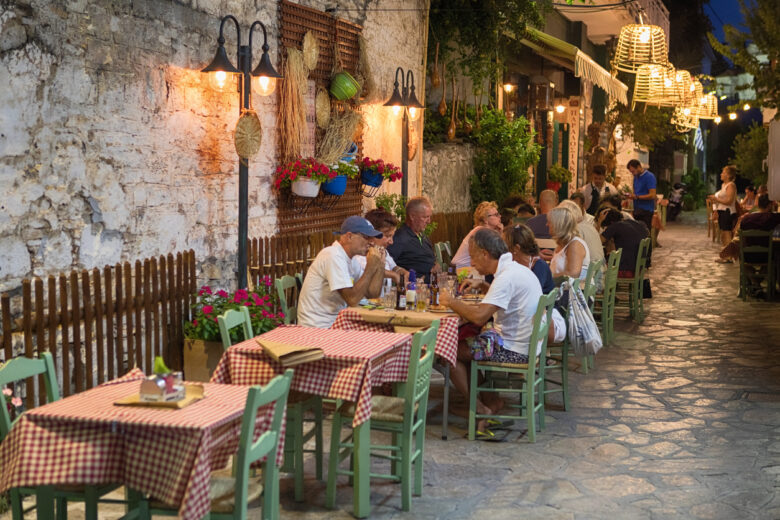 Pythagoreio is a small town and former municipality on the island of Samos, North Aegean, Greece. Greek cuisine is a Mediterranean cuisine. Contemporary Greek cookery makes wide use of vegetables, olive oil, grains, fish, wine, and meat. People in the Image are tourists enjoying the food.